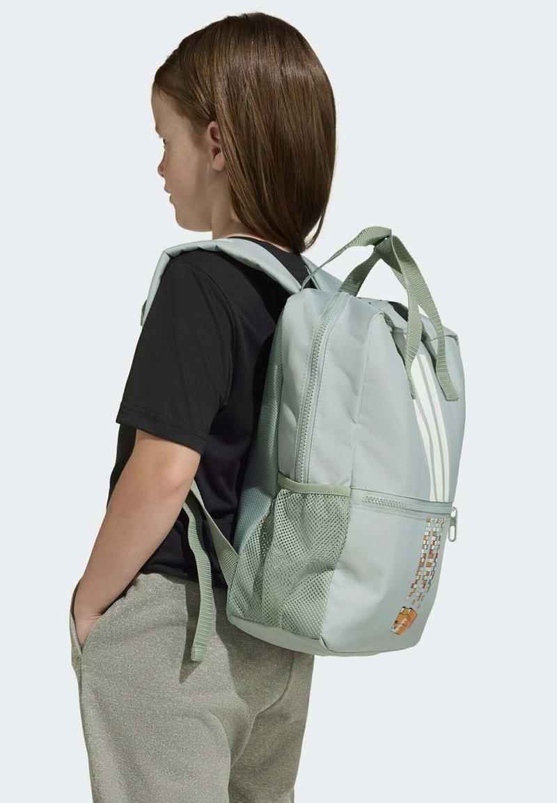 Zeleni ruksak s teksturiranim završetkom, s dvije mrežaste bočne džepove i tri bijele pruge. Model nosi crnu majicu i sive hlače.