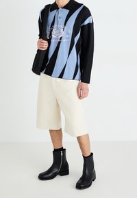 Polo shirt with blue and black wave pattern, buttoned collar, cream shorts, and black ankle boots with zippers, held black quilted bag.