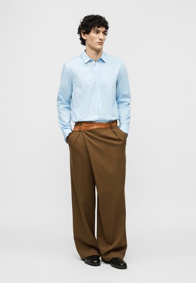 Un homme se tient les mains dans les poches, portant une chemise bleu clair boutonnée, un pantalon large marron avec une ceinture texturée, et des chaussures noires.