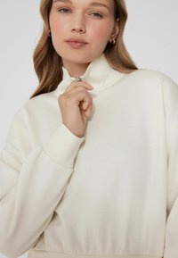 Kremowa bluza zapinana na zamek wykonana z miękkiego materiału, z wysokim kołnierzem, prążkowanymi mankietami i dopasowanym pasem o gładkiej fakturze.