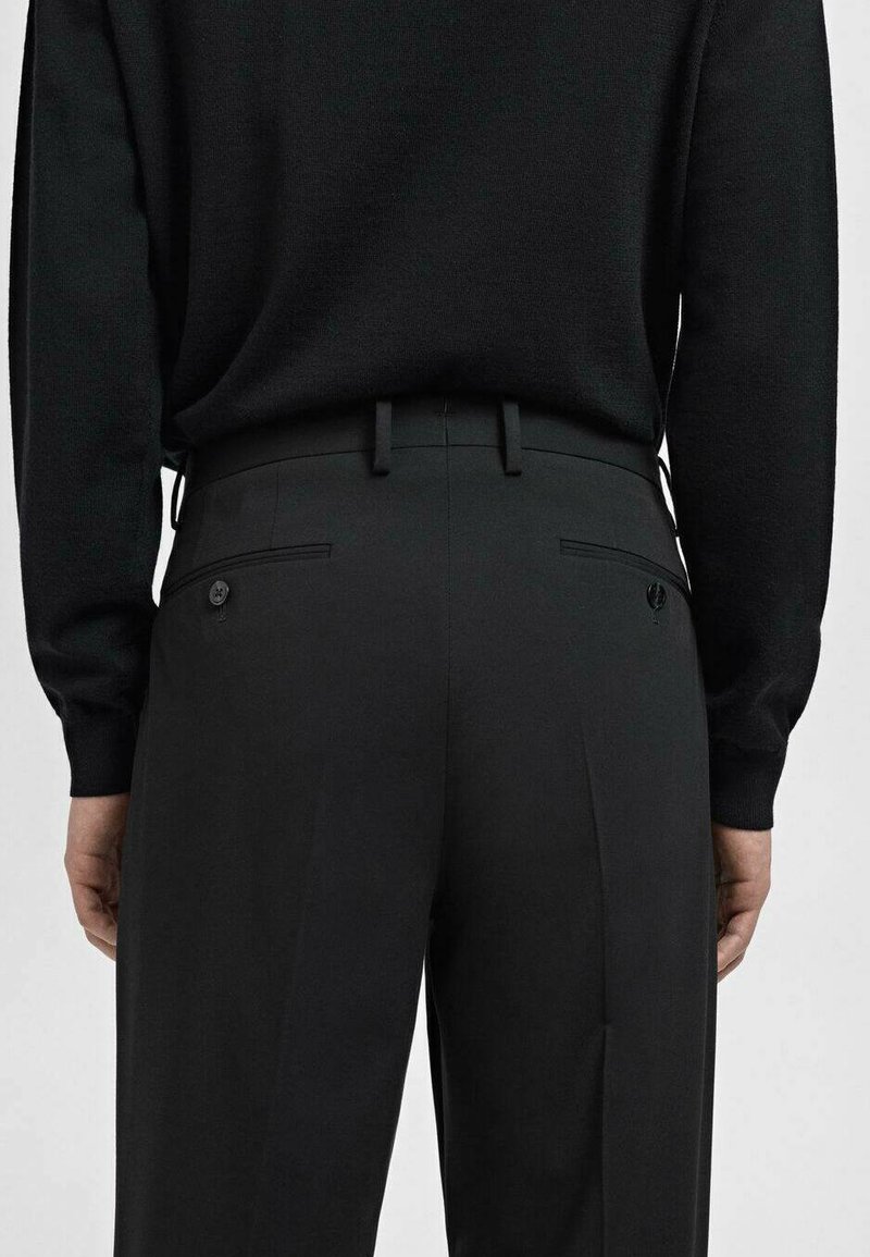 Pantalones negros de tejido suave, con un corte entallado, lazos para cinturón, bolsillos traseros con cierre de botón y una línea de planchado en cada pierna.