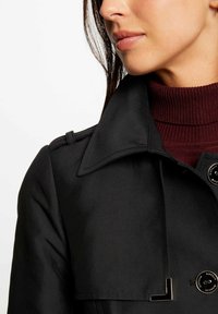 Zwarte trenchcoat met een brede kraag, zijknopen en een gladde textuur. Draagt over een bordeauxrode geribde coltrui.