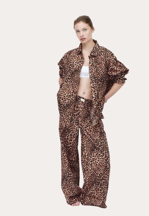 Kvinde iført løs leopardprint skjorte og bukser med en hvid cropped top, stående med hænderne i lommerne mod en ensfarvet baggrund.