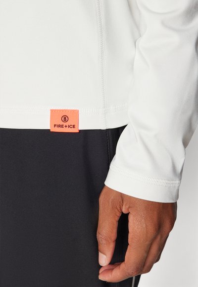 Λευκό πουκάμισο με μακριά μανίκια και ελαστικές μανσέτες, με μια πορτοκαλί ετικέτα λογότυπου στο στρίφωμα. Μαύρο παντελόνι με ομαλή υφασμάτινη υφή.