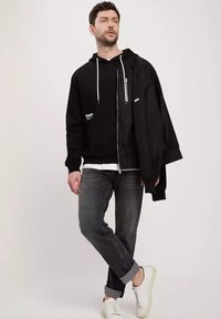 Czarna bluza z kapturem i białymi sznurkami oraz logo, nałożona na białą koszulę. Zestawiona z ciemnymi dżinsami i białymi sneakersami.