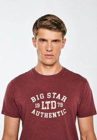 Vynio maroon spalvos medvilnės marškinėliai su apvalia iškirpte, papuošti baltais nusidėvėjusiais užrašais: "BIG STAR 19 LTD 79 AUTHENTIC." Trumpomis rankovėmis, laisvo silueto.