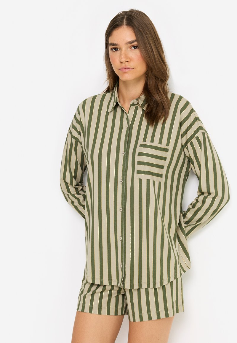 Femme aux cheveux bruns portant une chemise à boutons à rayures verticales vertes et beiges et un short assorti, debout devant un fond blanc.