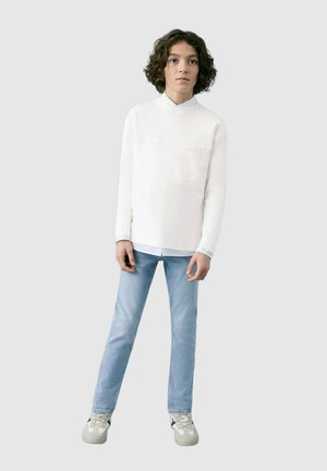 Valkea pitkähihainen paita, jossa on korkea kaulus ja rintatasku, yhdistetty vaaleansinisiin farkkuihin ja harmaisiin tennareihin, joissa on mustia yksityiskohtia.