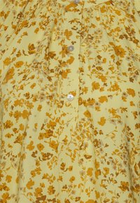 Tecido amarelo claro com um padrão floral em tons de castanho e mostarda, com uma casa de botões na frente. Textura suave.