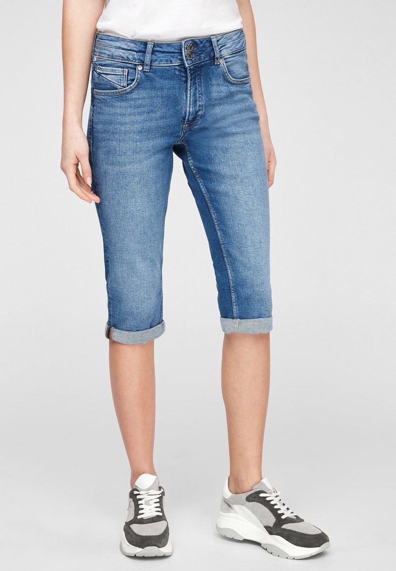 Pantalons capri en denim bleu clair avec ourlets roulés, design à cinq poches et fermeture à bouton en métal, associés à des baskets grises et blanches.