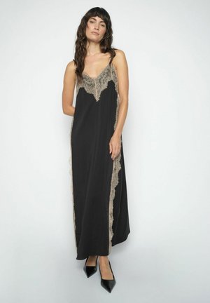 Kvinde står iført en sort ærmeløs maxikjole med beige blonder og sorte spidse hæle mod en hvid baggrund.