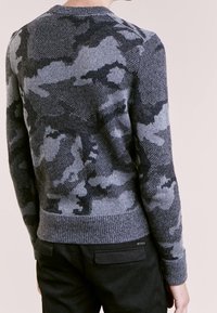 Grå stickad tröja med ett kamouflage-mönster, som har rund halsringning, ribbad nederkant och långa ärmar. Tyget är mjukt och texturerat.