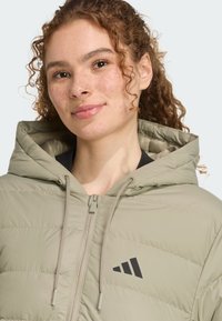 Ľahká, olivovo zelená prešívaná bunda s kapucňou, ktorá má predný zips, šnúrky na stiahnutie a čierne logo na hrudi.