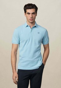 Polo shirt azzurro chiaro con colletto e maniche corte, caratterizzato da un piccolo logo "H" sul petto sinistro, abbinato a pantaloni scuri con texture.
