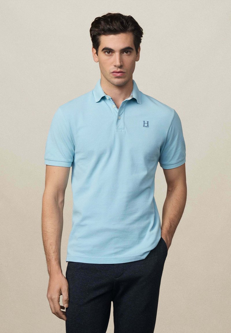 Polo shirt azzurro chiaro con colletto e maniche corte, caratterizzato da un piccolo logo "H" sul petto sinistro, abbinato a pantaloni scuri con texture.