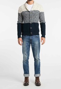 Cárdigan de punto con cuello de chal, en bloques de crema, gris y marino. Patrones texturizados y cinco botones, usado sobre unos vaqueros azules y botas marrones.