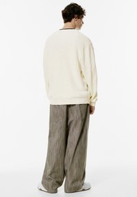 Pulover texturat de culoare crem, cu umeri lăsați, purtat împreună cu pantaloni largi gri cu dungi și adidași albi.