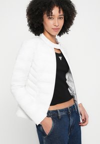 Fehér, enyhén puffos dzseki álló nyakkal, vízszintes varrással és cipzáras záródással, fekete testhez simuló felső és kék farmernadrág fölött viselve.