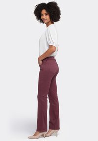 Jeans acampanados burdeos con bolsillos delanteros y traseros, combinados con una blusa blanca de mangas abullonadas. La modelo lleva tacones beige y está de perfil.