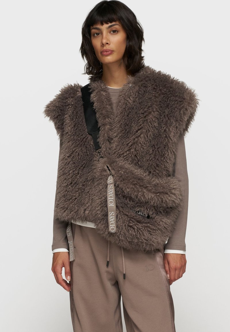 Brun faux fur vest med bred krave, lynlåslukning og justerbar rem. Sammen med beige bukser, der understreger tekstur og lagdeling.