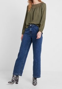 Blusa testurizzata verde oliva con scollatura quadrata, abbinata a jeans blu a vita alta con gamba dritta e stivaletti alla caviglia con stampa serpente bianca e nera.