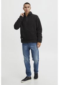 Sort, fluffy fleece-pullover med halv-zip-design og frontlomme, parret med blå jeans og sorte sneakers, sat mod en ensfarvet baggrund.