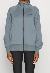 Grå zip-up jacka av mjukt material, har hög krage, raglanärmar och en logotyp på vänster bröst. Svarta accenter på dragkedjor.