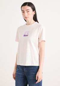 T-shirt in cotone rosa chiaro con maniche corte e vestibilità rilassata, con un logo quadrato metallicamente viola e oro CK sul petto.