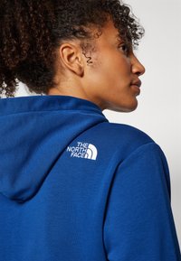 Modrá mikina s kapucňou vyrobená z jemného materiálu, s bielym logom "The North Face" na hornej časti chrbta. Dizajn obsahuje kapucňu so šnúrkou.