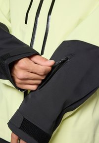 Veste imperméable jaune clair avec des accents noirs et une poche zippée sur le poignet gris foncé. Texture douce et coupe ajustable.