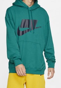 Turkoosipaita, joka on valmistettu pehmeästä kankaasta, ja jossa on suuri musta Nike-logo edessä, kengurutasku ja säädettävä nyörihuppu.