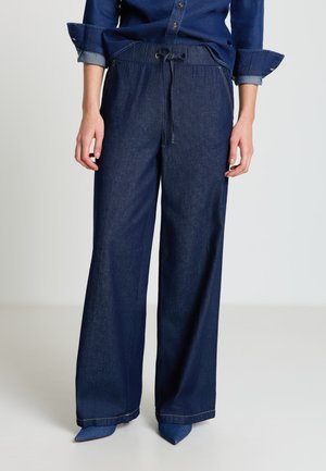 Persona che indossa pantaloni di jeans scuri a gamba larga con vita regolabile con coulisse e scarpe a punta abbinate in denim scuro, mani rilassate lungo i fianchi.