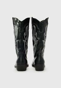 Botas de vaquero de cuero negro con costuras decorativas en forma de llama en plata, punta afilada y bordes superiores en forma de escote. Tacón bajo con suela texturizada.