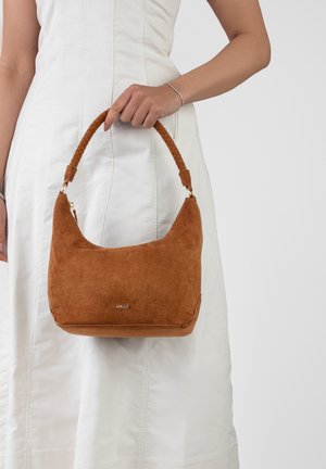 Γυναίκα με λευκό φόρεμα κρατά καφέ σουέτ τσάντα χειρός με πλεγμένη λαβή και χρυσά μεταλλικά στοιχεία, με την ετικέτα "RAID" στο κέντρο μπροστά.