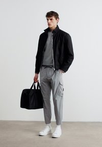 Νεαρός άνδρας που φοράει μαύρο μπουφάν, γκρι παντελόνι εργασίας, λευκά αθλητικά παπούτσια, κρατώντας μαύρη τσάντα duffel, στέκεται μπροστά σε απλό λευκό τοίχο.