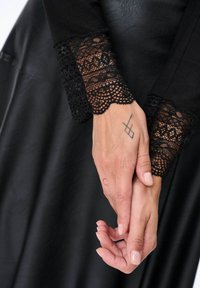 Czarny długi rękaw top z delikatnymi koronkowymi mankietami, noszony z czarną skórzaną spódnicą. Ręce złożone, z widocznym tatuażem i naturalnymi paznokciami.