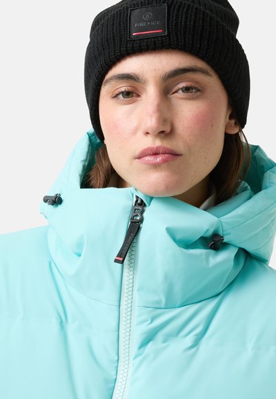 Jasno-niebieska kurtka puchowa z wysokim kołnierzem i zamkiem błyskawicznym. Czarna dzianinowa czapka z daszkiem na głowie, z naszywką z logo. Gładka faktura.