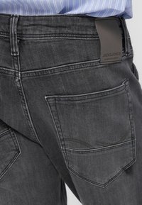 Σκούρο γκρι τζιν με αντίθετες ραφές, με ετικέτα της μάρκας στη μέση και δύο πίσω τσέπες με στρογγυλεμένες άκρες.