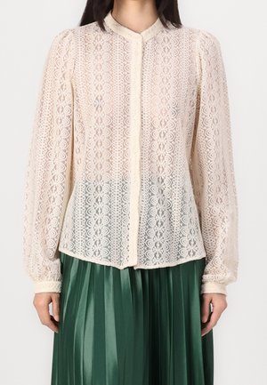 Blouse blanche en dentelle à manches longues avec motif floral et boutons sur le devant, portée avec une jupe plissée vert foncé.