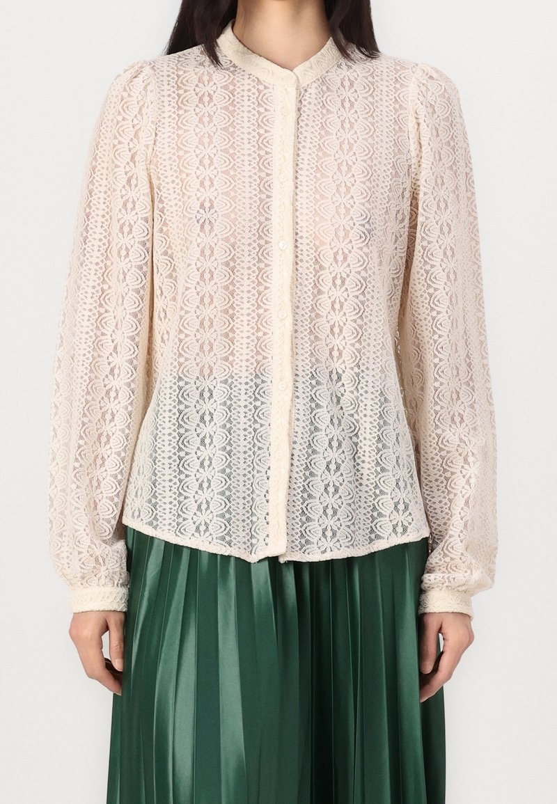 Blouse blanche en dentelle à manches longues avec motif floral et boutons sur le devant, portée avec une jupe plissée vert foncé.