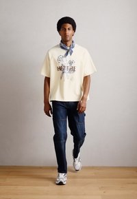 Hvid T-shirt med korte ærmer og blomster grafik, parret med blå denim jeans og hvide sneakers. Modellen bærer et blåt tørklæde om halsen.