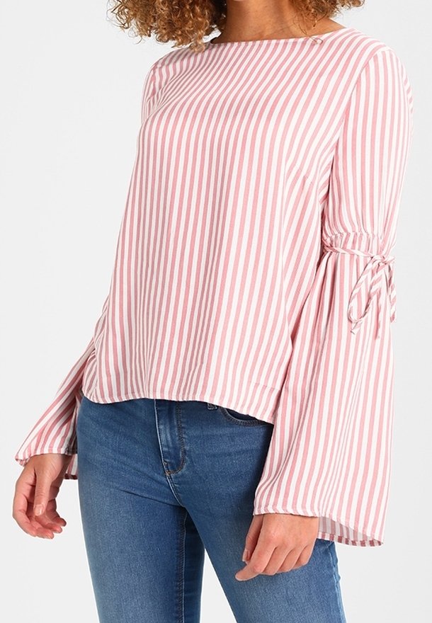 Femme portant un chemisier à manches longues rayé verticalement rose et blanc avec des manches évasées nouées aux coudes, associé à un jean bleu.