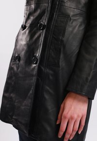 Cappotto di pelle nera con design doppiopetto, quattro grandi bottoni, tasca laterale e texture liscia. Parte del braccio visibile.
