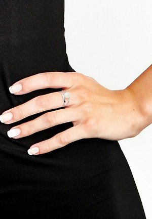 Zilveren ring met een kleine witte parel, met een minimalistisch ontwerp. De ring heeft twee in elkaar grijpende banden en wordt gedragen aan een hand met gepolijste nagels.