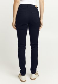 Jeans ajustados de color azul oscuro con cintura alta, diseño de cinco bolsillos y una etiqueta de piel en la parte trasera de la cintura, combinados con zapatillas de color claro.