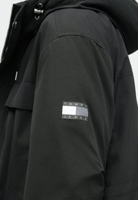 Camisola preta feita de um tecido suave e leve, apresentando um adesivo de logotipo na manga e um capuz com cordões ajustáveis.