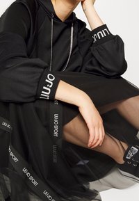 Czarna bluza z kapturem i długimi rękawami o fakturowanej powierzchni, noszona na przezroczystej czarnej spódnicy z logo "LIUJO SPORT".