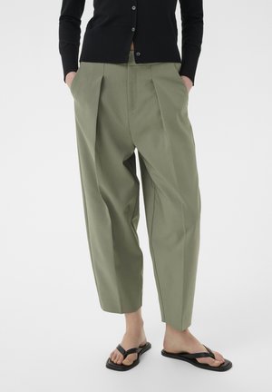 Olívazöld pliszírozott, bőszárú nadrágot, fekete gombos felsőt és fekete papucsot viselő személy áll fehér háttér előtt, kezét zsebében tartva.