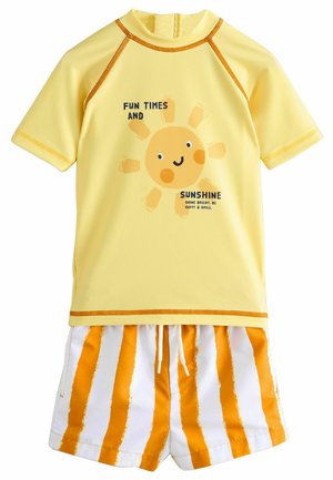 Gelbes Rash Guard mit kurzen Ärmeln, versehen mit einem lächelnden Sonnenmotiv und Text. Passende Badehose mit vertikalen Streifen in Orange und Weiß.