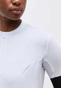 Κοντινό πλάνο ενός ατόμου που φοράει εφαρμοστό ανοιχτό γκρι πουκάμισο με φερμουάρ στο μπροστινό μέρος και πλέγμα κάτω από τη μασχάλη, μπροστά σε απλό φόντο.